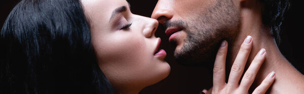 side view of sexy brunette woman kissing young man isolated on black, banner