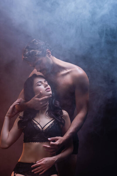 sexy shirtless man hugging neck of seductive woman in black lace lingerie on dark background with smoke