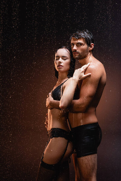 shirtless muscular man looking at camera while hugging sexy woman under rain on dark background