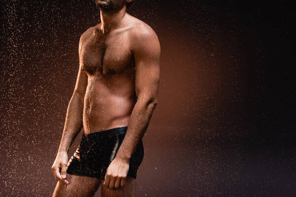 cropped view of sexy, muscular man in black underpants under falling raindrops on dark background