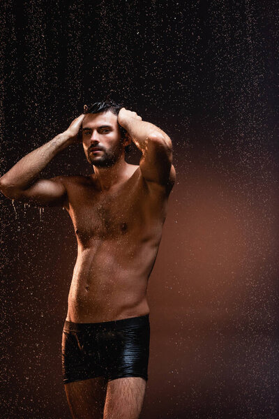 muscular shirtless man in black underpants touching head under rain on dark background