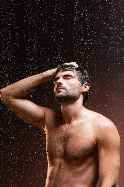 shirtless muscular man with closed eyes under falling raindrops on dark background