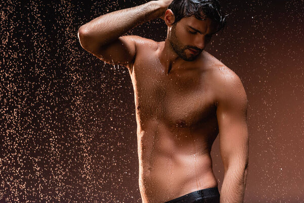 shirtless man with muscular torso posing with hand on head under rain on dark background