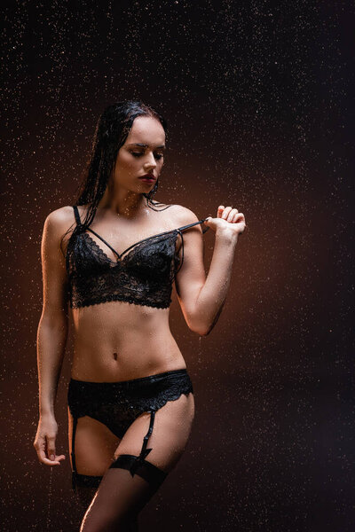 young woman in black lace underwear touching strip of bra under falling rain on dark background