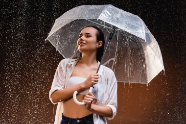 Sırıtan ıslak tişörtlü, şeffaf şemsiyeli, yağmurun altında koyu arka planda gülümseyen kadın.