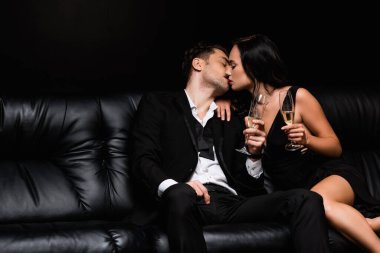 Seksi çift öpüşüyor ve şampanya ile bardaklarını tutarken siyah koltukta yalnız oturuyorlar. 