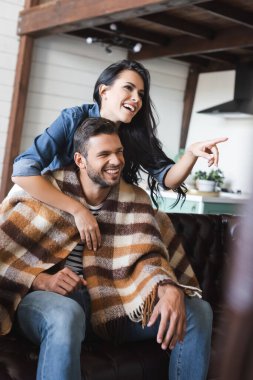 Kareli battaniyenin altında film seyreden bir adamın omzunu tutarken parmaklarıyla işaret eden neşeli kadın.