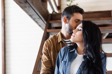 Genç adam, ahşap merdivenlerde gözleri kapalı esmer kız arkadaşını öpüyor.