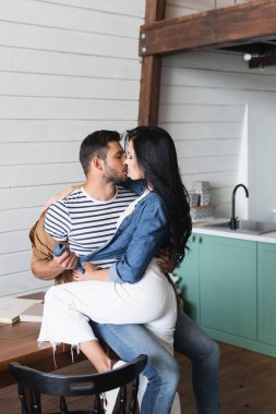Şık giyinmiş genç bir çift mutfakta sarılıp öpüşüyor.