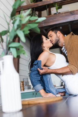 Genç adam soyunuyor ve seksi kadını öpüyor. Bulanık ön planda masada oturuyor.