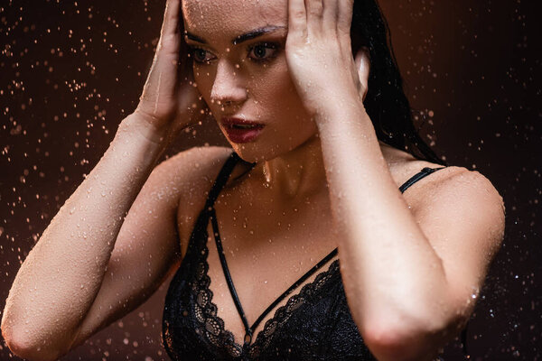 sensual woman in black lace bra posing with hands near face under rain on dark background