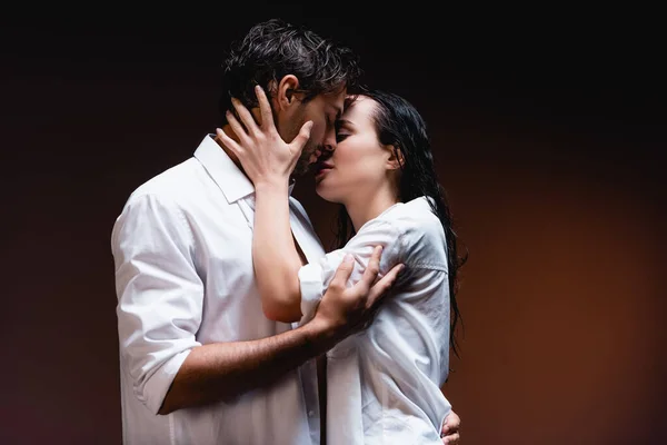 young couple in wet white shirts embracing and kissing on dark background
