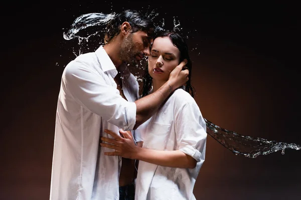 young couple in wet white shirts embracing with closed eyes near water splashes on dark background
