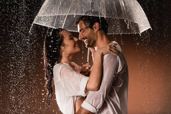 side view of happy couple embracing while standing with umbrella under rain on dark background