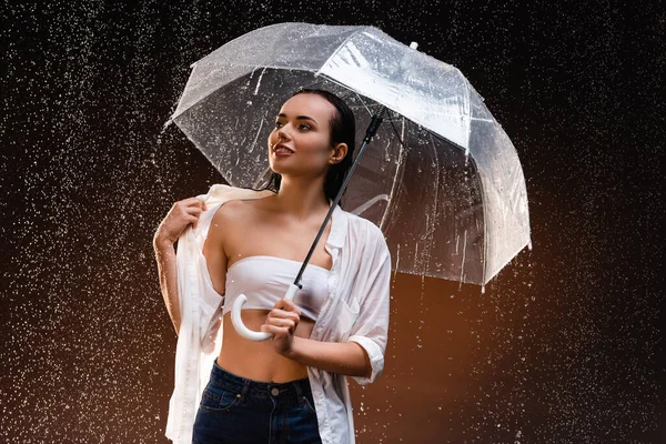 wet, sexy woman looking away while standing with umbrella under rain on dark background