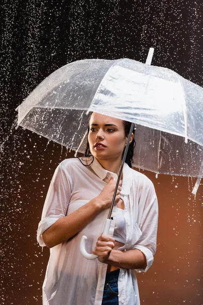 young woman in wet white shirt standing under rain with transparent umbrella on dark background