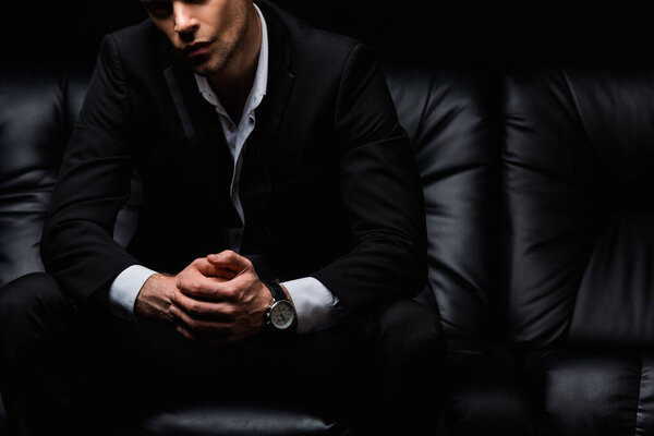 cropped view of confident man in suit sitting on black leather sofa 