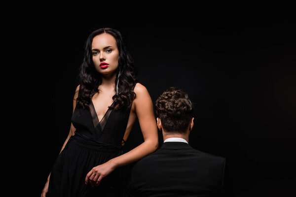 соблазнительная женщина в платье глядя на камеру рядом с мужчиной в костюме, изолированном на черном 