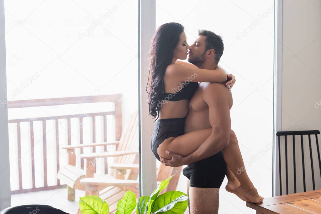 side view of sexy muscular man holding seductive woman in black lingerie against balcony window