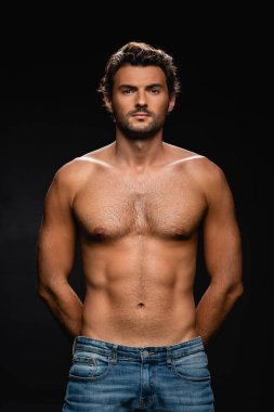 Siyah arka planda elleri arkada poz verirken kameraya bakan seksi üstsüz adam.