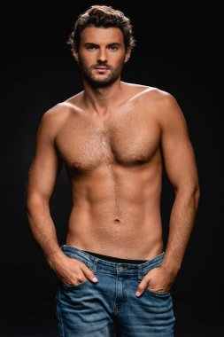 Siyah arka plandaki kameraya bakarken elleri ceplerinde poz veren kaslı, kendine güvenen bir adam.