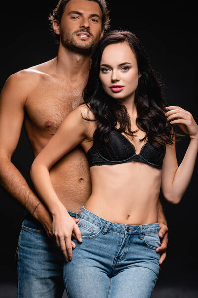сексуальный мускулистый мужчина и соблазнительная брюнетка женщина в лифчике и джинсах, смотрящая на камеру, изолированную на черном