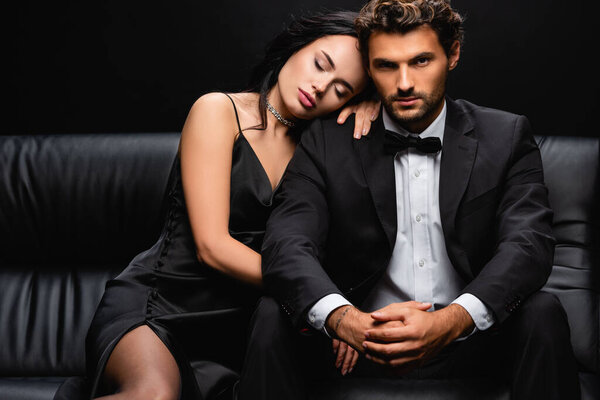 sexy woman in satin dress lying on shoulder of elegant man sitting on leather couch isolated on black