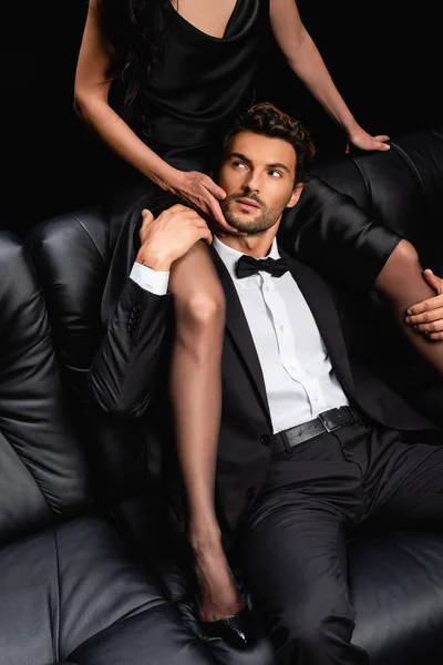 スーツを着た男の上のソファに座って黒で隔離された顔に触れるセクシーな女性 — ストック写真