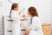Smiling mother and child in bathrobes brushing teeth in bathroom 