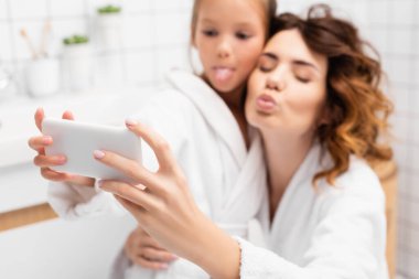 Anne ve çocuğun ellerinde akıllı telefon banyodaki bulanık arka planda selfie için poz veriyor. 