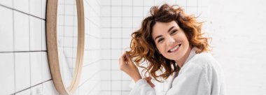 Cheerful woman in white bathrobe adjusting hair near mirror in bathroom, banner  clipart