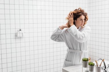 Woman in bathrobe and hands near hair looking away in bathroom  clipart