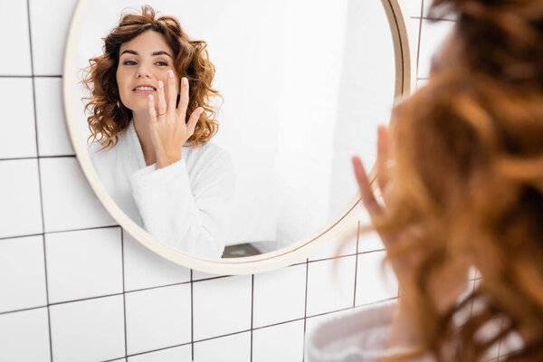 Smiling woman in bathrobe applying face cream near mirror on blurred foreground 