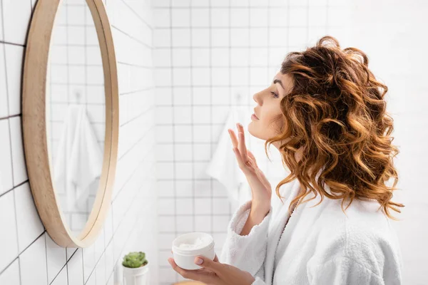 浴室靠近镜子的卷曲妇女使用面霜的侧视图 — 图库照片