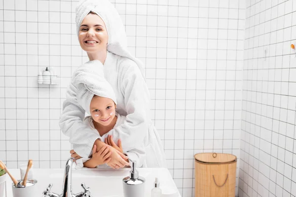 Donna Sorridente Asciugamano Accappatoio Abbracciando Figlia Con Crema Sul Viso — Foto Stock