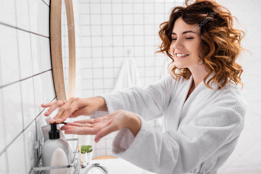 Cheerful woman applying cosmetic cream on blurred foreground in bathroom 