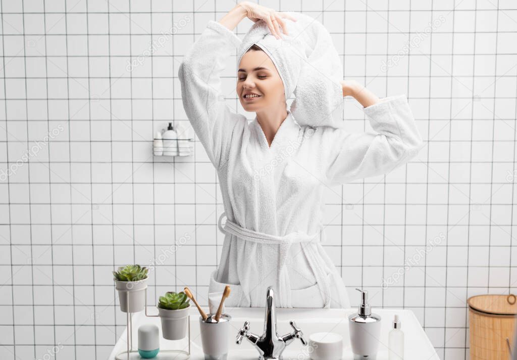 Positive woman in bathrobe and towel smiling with closed eyes near sink in bathroom 
