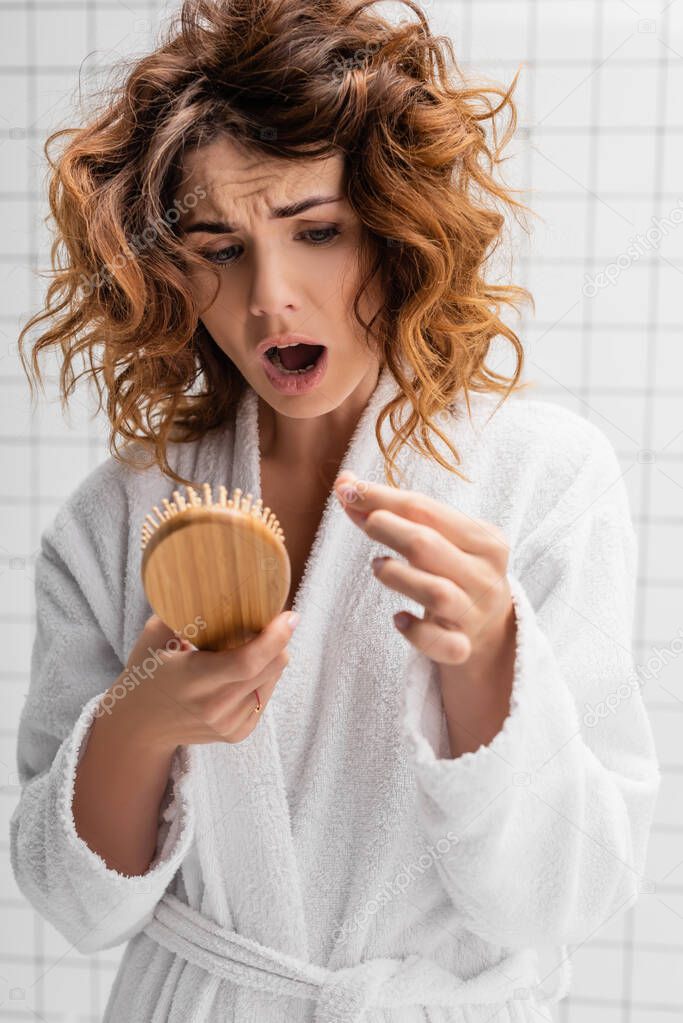 Displeased woman holding hair brush on blurred foreground in bathroom 