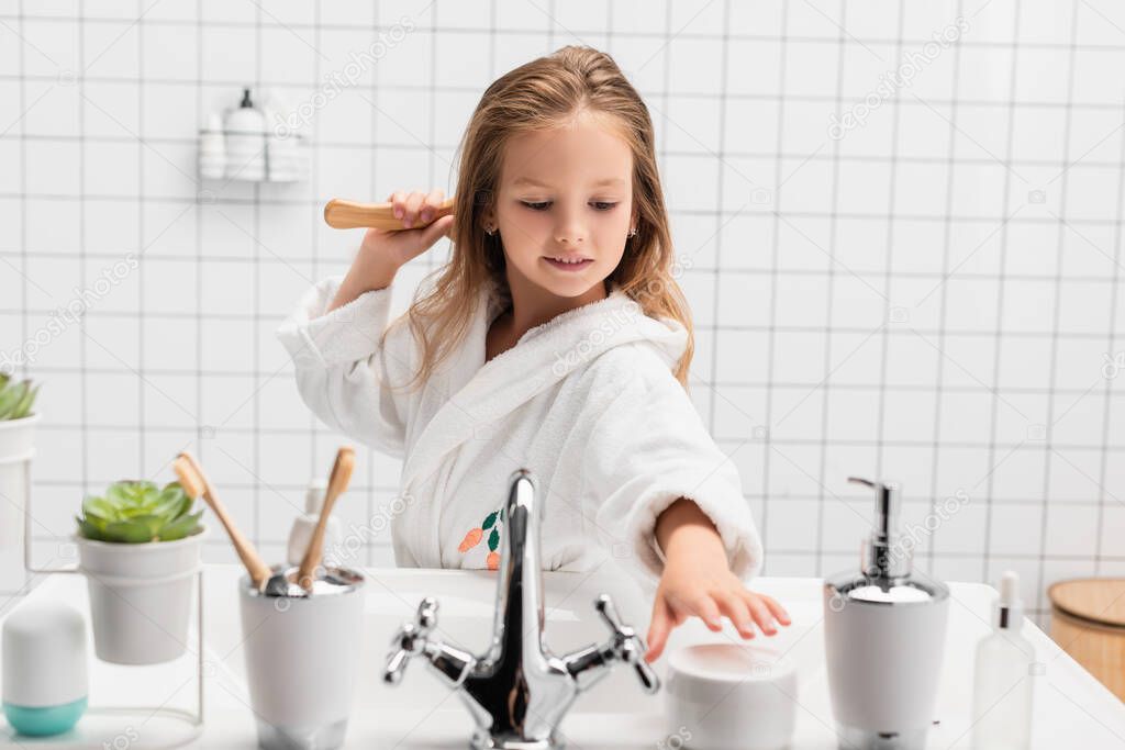 Smiling girl brushing hair and taking cream in bathroom 