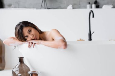 Köpüklü genç kadın evde banyo yaparken kameraya bakıyor. 