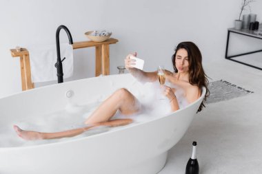 Genç bir kadın elinde bir bardak şampanyayla akıllı telefonuyla selfie çekiyor. 