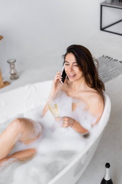 Bir bardak şampanyası olan neşeli bir kadın küvette köpüklü akıllı telefonuyla konuşuyor. 