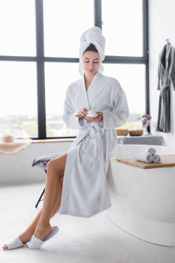 Modern banyoda banyo küvetinin yanında elinde kozmetik krem olan bornoz giymiş bir kadın. 