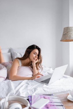 Akıllı telefonun yanında dizüstü bilgisayar ve yatak odasında bulanık ön plan kulaklık kullanan genç bir kadın. 