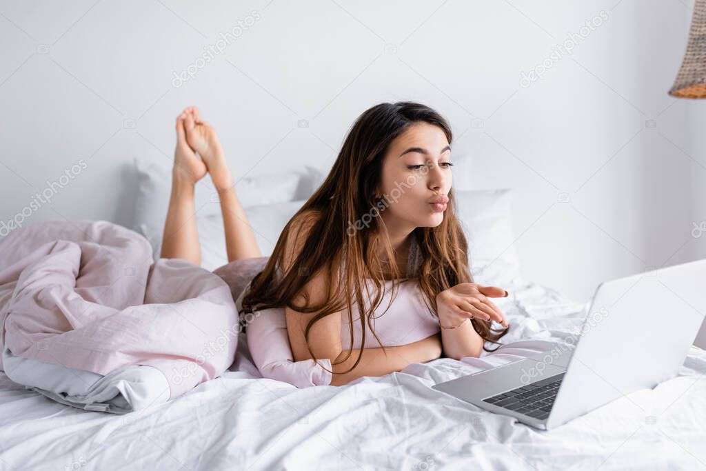 Woman blowing air kiss at laptop on blurred foreground on bed 