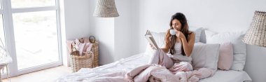 Young woman with cup of coffee reading book on bed, banner  clipart
