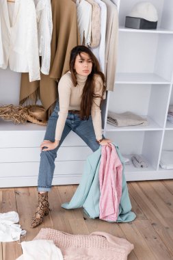 Pensive woman holding clothes while sitting on shelf in wardrobe  clipart