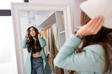 Young woman wearing hat while looking at mirror on blurred foreground in wardrobe  clipart