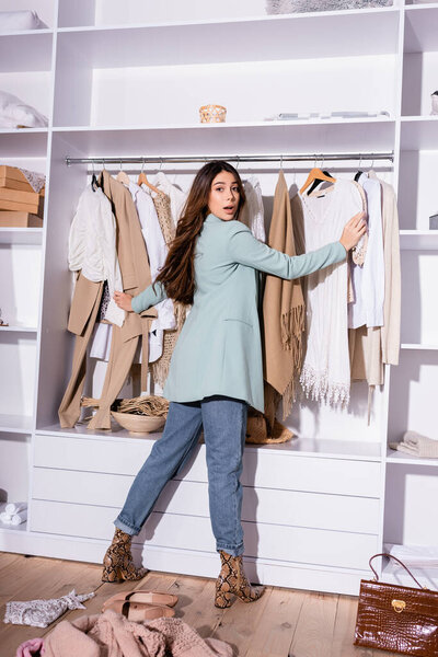 Back view of woman looking at camera while standing near clothes in wardrobe 