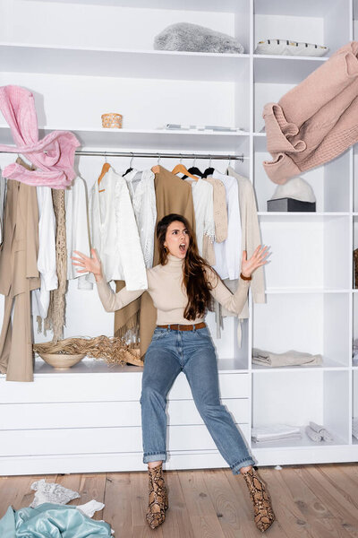 Angry woman throwing clothes while sitting on shelf in wardrobe 